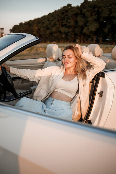 Czarująca luksusowa dziewczyna z blond włosami uśmiechnięta siedząc na białym kabriolecie. Młoda kobieta sukcesu siedzi w swoim białym samochodzie - Zdjęcie, obraz