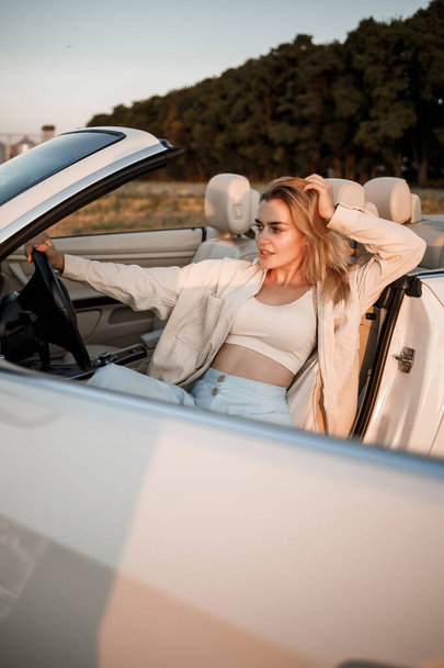 Lumoava luksustyttö, jolla on vaaleat hiukset ja joka hymyilee istuessaan valkoisella avoautolla. Nuori menestynyt nainen istuu hänen valkoinen auto - Valokuva, kuva