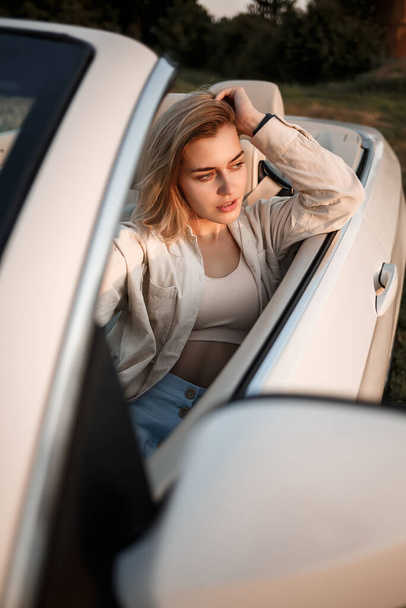 A glamorous luxury girl with blonde hair smiling while sitting on a white convertible. Young successful woman sitting in her white car - Foto, Bild