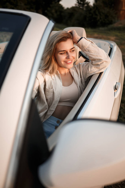 A glamorous luxury girl with blonde hair smiling while sitting on a white convertible. Young successful woman sitting in her white car - Foto, imagen