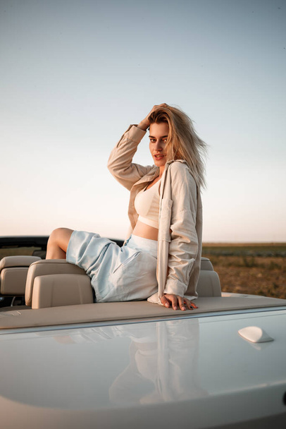 Okouzlující luxusní dívka s blonďatými vlasy, usmívající se, zatímco sedí na bílém kabrioletu. Mladá úspěšná žena sedí ve svém bílém autě - Fotografie, Obrázek