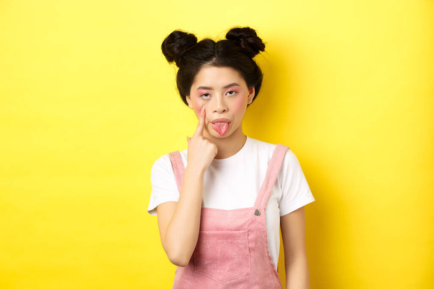 Грубая азиатская девушка растягивает веко и показывает язык, насмехаясь над кем-то, стоя на желтом фоне - Фото, изображение