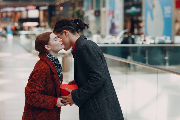 Un tipo le da a su novia una caja en forma de corazón en el Día de San Valentín. Pareja joven besándose en el centro comercial. - Foto, imagen
