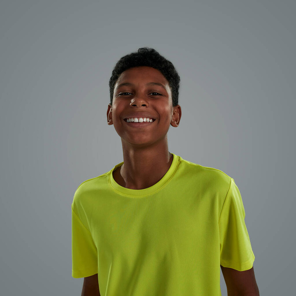 Young sportsman, Portrait of a happy cheerful teenage african boy wearing neon t shirt smiling at camera while standing against grey background in studio - Фото, изображение