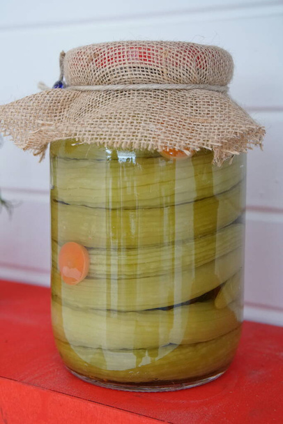 Komkommers in blik met specerijen in glazen pot. Glazen pot met gepekelde komkommers op rode houten plank - Foto, afbeelding