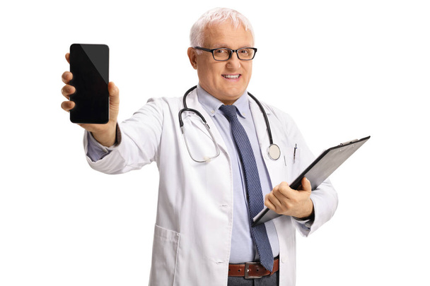 Mature male doctor holding a clipboard and showing a mobile phone isolated on white background - Zdjęcie, obraz