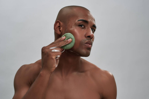 Portrait of handsome african american young man enjoying skincare procedure, using cleansing sponge while washing his face isolated over gray background - Fotó, kép