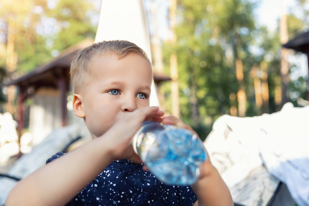 Portret van schattig speels klein blank blond dorstig peuter jongen met plastic fles en drinkwater op hete zomerdag buiten. Kinderen verwarmen uitdroging gevaar bewustzijn - Foto, afbeelding