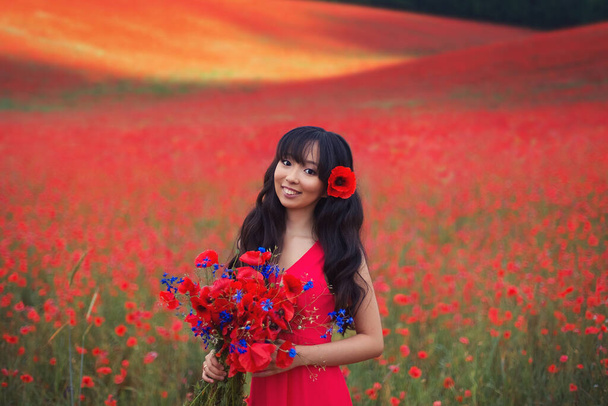 Gyönyörű ázsiai lány hosszú haj egy piros ruhát egy nagy csokor mák és búzavirág a kezében a háttérben egy virágzó mező vad vörös virágok. - Fotó, kép