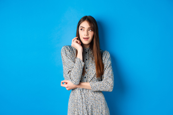 Pensive caucasian girl with long natural hair, wearing vintage dress, looking left with thoughtful face, standing on blue background - Photo, Image