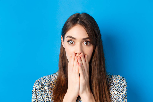 People emotions concept. Shocked young woman face, covering mouth with hands and gasping, look startled, standing on blue background - Photo, Image