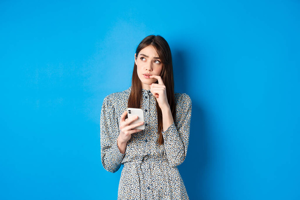 Pensive söpö tyttö ajattelu miten vastata viestiin, katsomatta sivuun harkittuja ja tilalla älypuhelin, seisoo mekko sinisellä taustalla - Valokuva, kuva