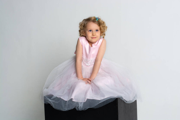 ένα μικρό κορίτσι σε ένα ροζ φόρεμα και στέμμα κάθεται σε ένα μαύρο κύβο σε λευκό φόντο - Φωτογραφία, εικόνα