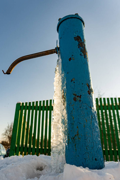 Blauwe waterpomp bedekt met ijs, het water bevroor op de drinkwaterkolom, ijs uit de waterput - Foto, afbeelding