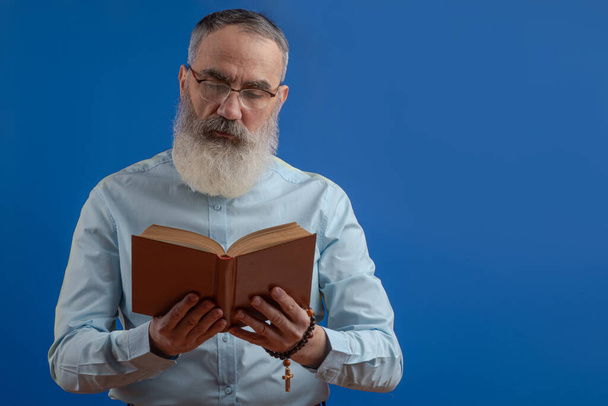 Homem carismático sênior com uma barba cinza em uma camisa clara lendo um livro A Bíblia Sagrada, rosário com uma cruz em um fundo azul isolado - Foto, Imagem