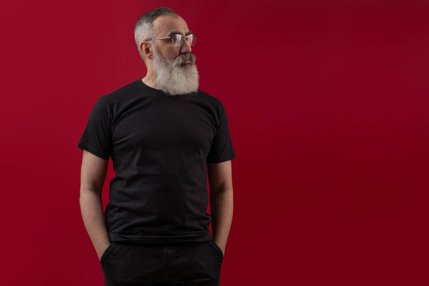 Adult handsome senior male model with a gray beard wearing a black t-shirt. Front view. Mock up space for your logo or design over dark red isolated background - Valokuva, kuva