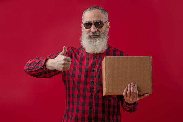 Dospělý pohledný starší muž s šedým plnovousem v kostkované červené a černé košili, s palcem nahoru, vypadá sebevědomě a v ruce drží lepenkovou krabici. Vysmívej se. Červené izolované pozadí - Fotografie, Obrázek