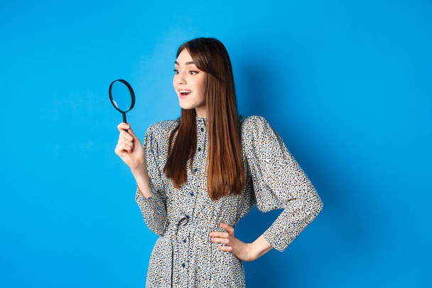 Excited girl looking left with magnifying glass, found interesting promo, investigating or searching, standing on blue background - Photo, Image