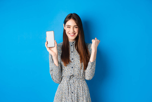 Cheerful pretty girl chanting, winning on mobile phone, showing empty smartphone screen and fist pump, smiling and celebrating, blue background - Photo, Image