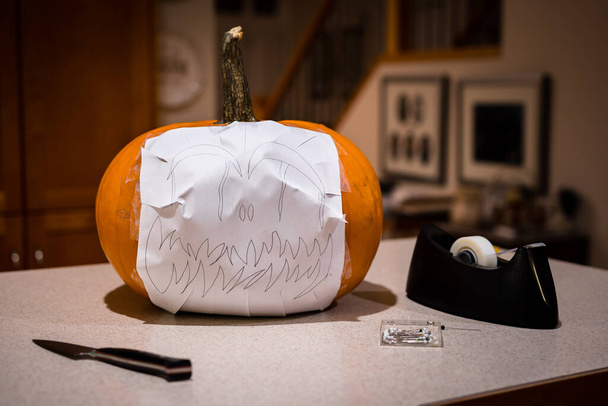 Φτιάχνω ένα Jack-o-lantern για το Halloween στην κουζίνα. Πώς να χαράξετε μια κολοκύθα χρησιμοποιώντας ένα προκατασκευασμένο πρότυπο σχεδιασμού. Δημιουργικό σχέδιο χαρτί ακολουθήσει κατά μήκος μοτίβο και μαχαίρι από το σπίτι - Φωτογραφία, εικόνα