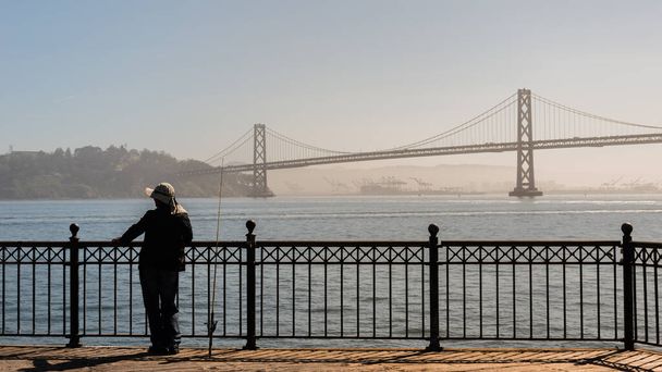 サンフランシスコの桟橋で釣りをしている人。オークランドベイブリッジで水平線の煙と煙。悪天候や山火事による大気汚染 - 写真・画像