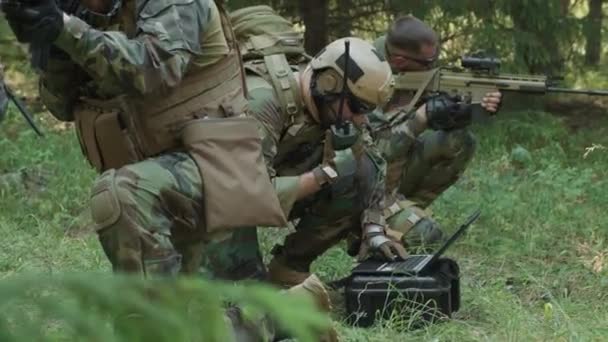 Medium shot of squad of special forces soldiers on military operation in jungle, commander using radio while transmitting data on laptop, and others covering him with rifles - Footage, Video