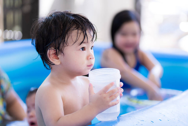 A kisfiú szomjan akart inni egy kis vizet. A gyerekek kék gumi medencében játszanak. Nyáron vagy tavasszal. A gyerek szabadidős tevékenységet folytat a családjával a ház előtt. 1-2 éves fiú. - Fotó, kép
