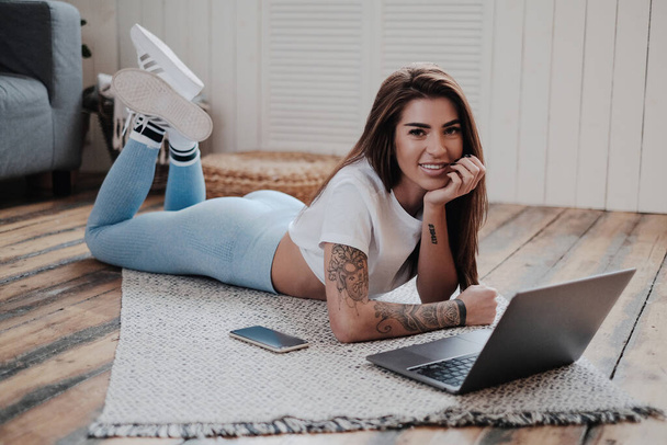 Optimistisch en stijlvol meisje liggend op de vloer met moderne laptop in de kamer - Foto, afbeelding