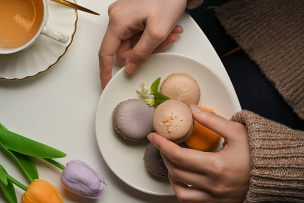 Vista dall'alto della mano femminile raccogliendo macaron da mangiare con tè sul tavolino decorato con fiori di tulipani - Foto, immagini