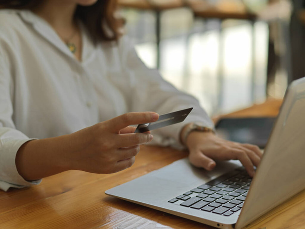 Vista lateral de la mano femenina que sostiene la tarjeta de crédito mientras está en línea pagando en el ordenador portátil  - Foto, imagen