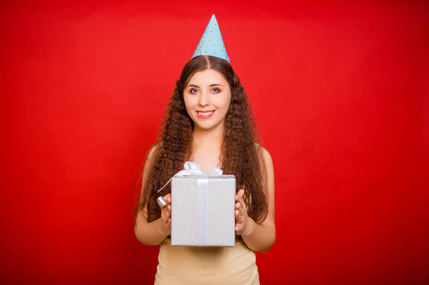 Portrait of an attractive girl in a festive cap with a gift in her hands and blowing a pipe isolated on red background. Happy and noisy birthday concept. - Foto, immagini