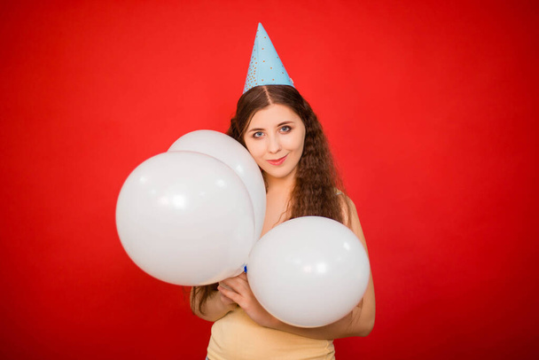 Portrait of an attractive young woman in a festive cap with white balloons in her hands, isolated on a red background. Happy birthday concept. - Foto, immagini