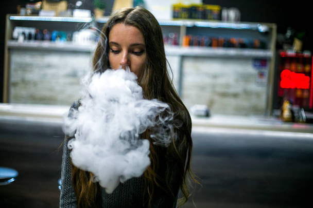 Vaping teenager. Young pretty white caucasian teenage girl with long hair smoking an electronic cigarette indoors. Deadly bad habit. Vape activity. - Фото, изображение
