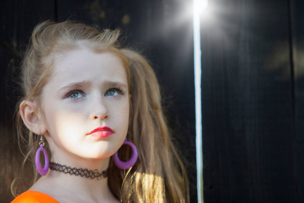 Parlak makyajlı küçük bir kızın portresi ahşap bir çitin yanında duruyor. Kızıl saçlı küçük kız.. - Fotoğraf, Görsel