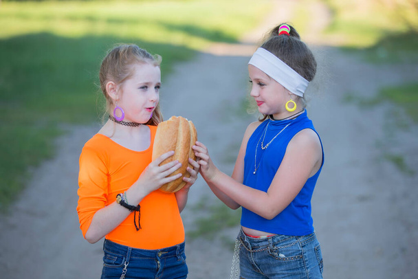 Dos chicas con maquillaje brillante vestidas al estilo de los noventa se están comiendo un bollo. Aldea rusa niños. - Foto, imagen