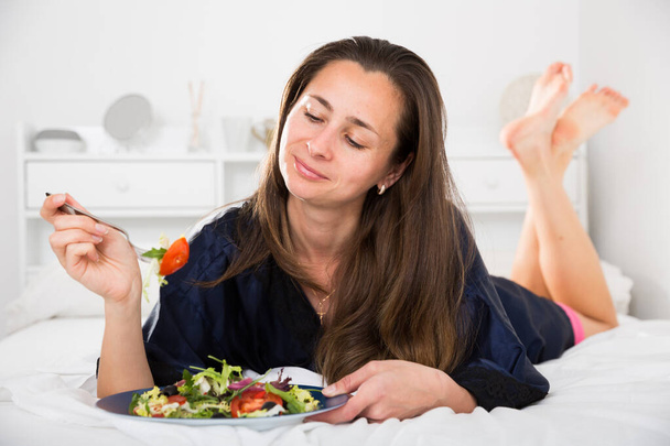 Slim female eating salad in bed at home and smiling - Fotó, kép