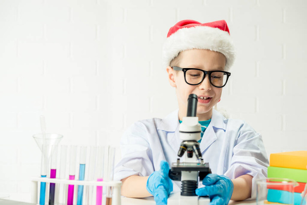 Ученик изучает химию. Позитивное радостное тестирование умных мальчиков и использование микроскопа на уроке естественных наук в шляпе Санты - Фото, изображение