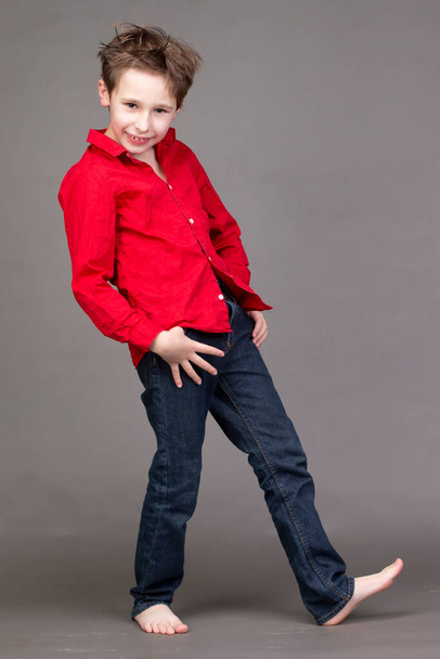 Красавчик в красной рубашке и джинсах на сером фоне. Ребенок в модельном бизнесе позирует. - Фото, изображение