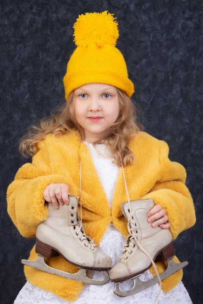 Piękna dziewczynka w zimowych ubraniach z białymi łyżwami. Pięcioletnie dziecko w dzianym żółtym kapeluszu. - Zdjęcie, obraz