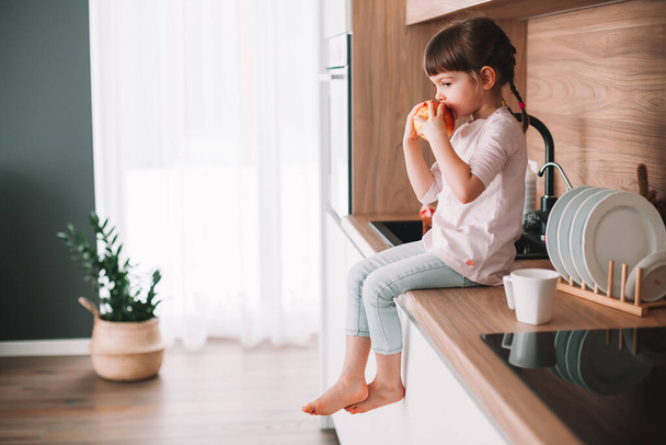 Cute little girl jedzenia czerwone jabłko siedzi na powierzchni kuchni. Koncepcja zdrowego odżywiania. - Zdjęcie, obraz