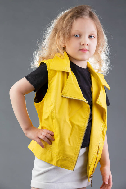 Krásná malá blondýnka ve žluté bundě na šedém pozadí. Pětiletá holka. Testy modelu pro dítě. - Fotografie, Obrázek