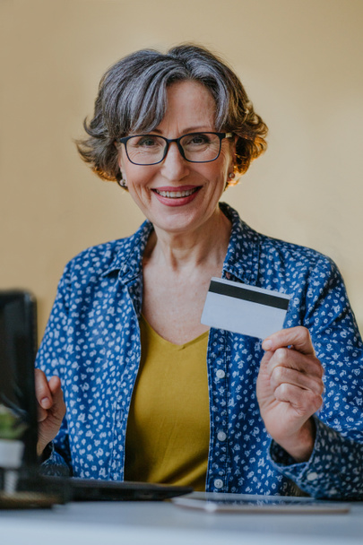 Retrato de mulher elegante feliz sênior com cabelos grisalhos fazendo shooping online usando laptop com cartão de crédito indoor. - Foto, Imagem