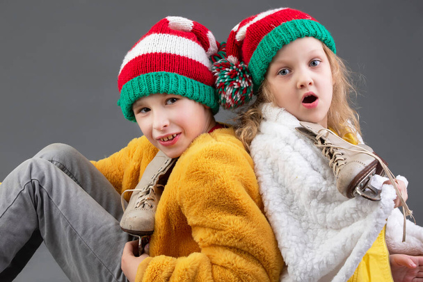Muchacha divertida y niño en sombreros de Navidad de punto y patines de hielo vintage se sientan con la espalda el uno al otro sobre un fondo gris. Feliz Navidad niños. - Foto, imagen