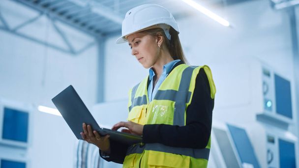 High-Tech Factory: Confident and Professional Female Engineer Wearing Safety Jacket and Hard Hat Holding and Working on Laptop Computer. Modern Bright Industrial Facility. Low Angle Shot - Фото, изображение