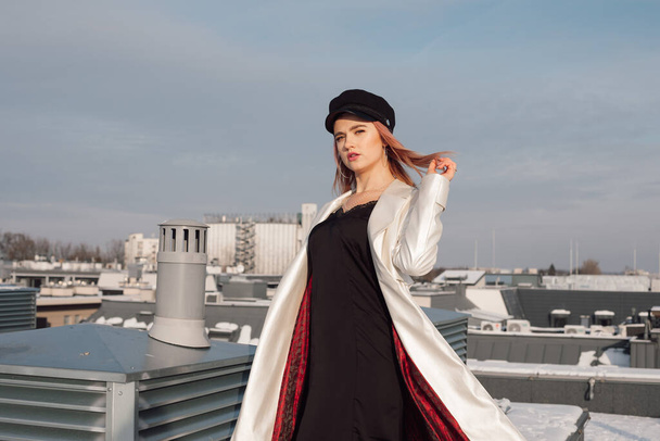 Žena na střeše budovy proti modré obloze v paprscích chladného zimního slunce. Má na sobě černé teplákové šaty, bílý plášť s červenou podšívkou a čepici. Drží její zrzavé vlasy ve větru - Fotografie, Obrázek