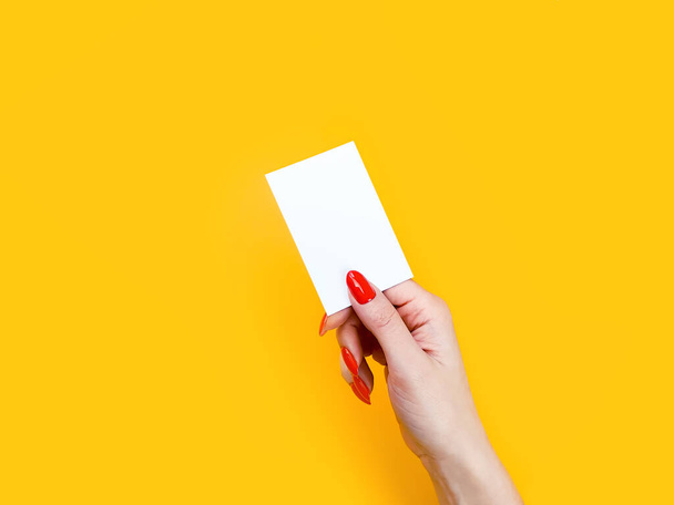 Female hand with red manicure gives blank white business card, close-up, isolated on yellow background. Mockup - Фото, изображение