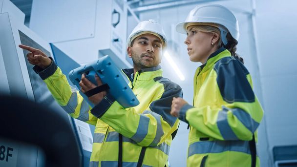 Chief Engineer and Project Manager Wearing Safety Vests and Hard Hats, Use Digital Tablet Controller in Modern Factory, Talking, Programming Machine For Productivity. Low Angle Portraits - 写真・画像