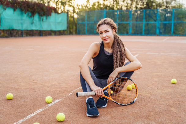 Портрет красивой молодой женщины, сидящей рядом с теннисным кортом среди теннисных мячей на открытом воздухе. Спортсменка отдыхает на теннисном корте. Позирование в стильной спортивной одежде. - Фото, изображение