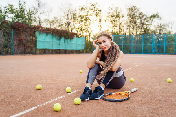 Portrait of a beautiful young woman sitting near a net tennis court among tennis balls outdoors. Sportswoman resting on a tennis court. Posing dressed in stylish sportswear. - Photo, Image