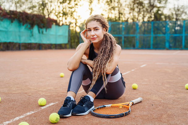 Porträt einer schönen jungen Frau, die in der Nähe eines Netztennisplatzes inmitten von Tennisbällen im Freien sitzt. Sportlerin ruht sich auf einem Tennisplatz aus. Posieren in stylischer Sportkleidung. - Foto, Bild